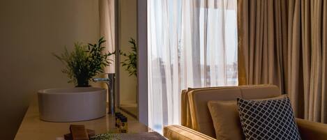 A modern hotel room with a sofa, a round white side table, a potted plant, and a window with sheer curtains.