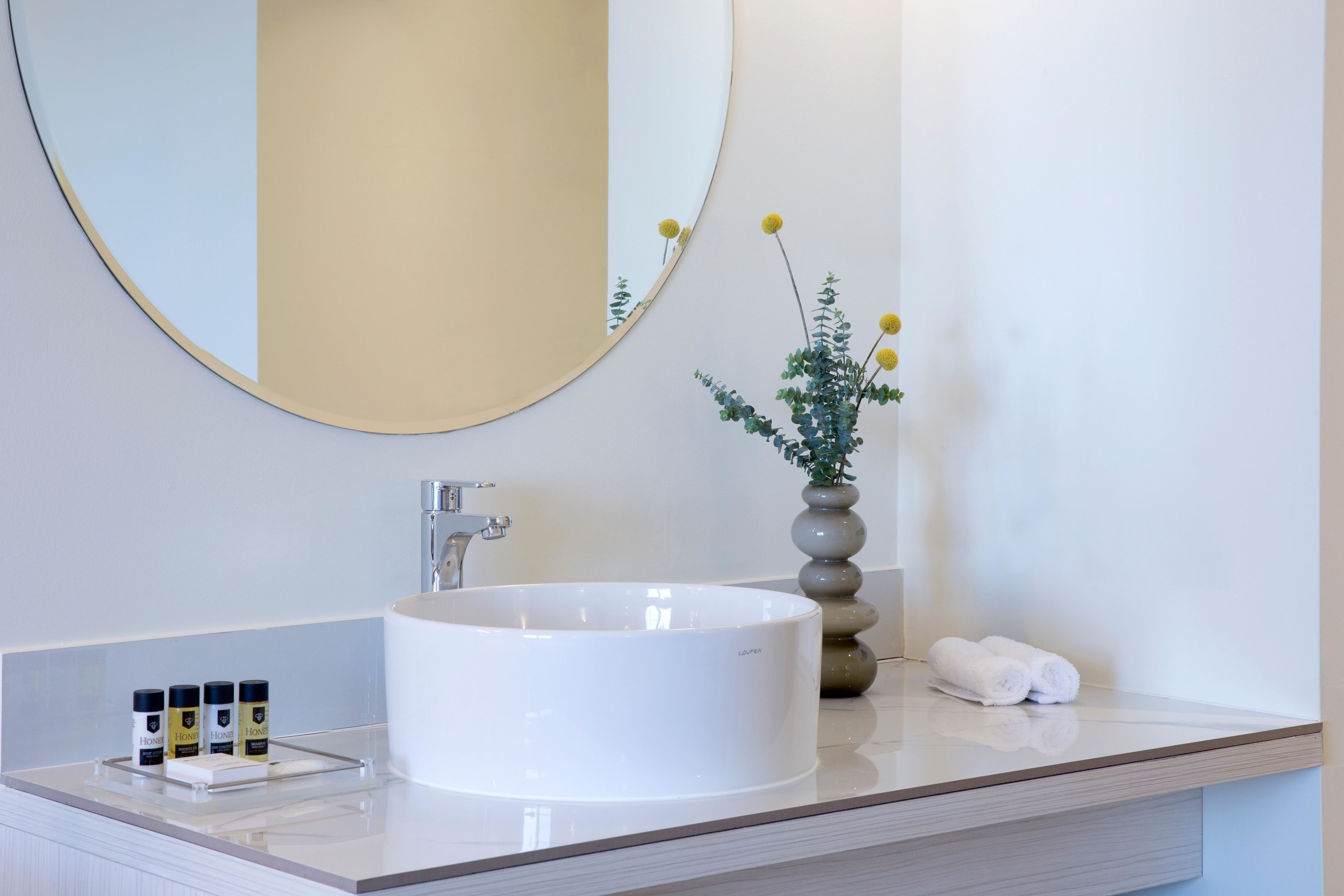 A modern bathroom with a round white sink, a large mirror, and a vase with yellow flowers.