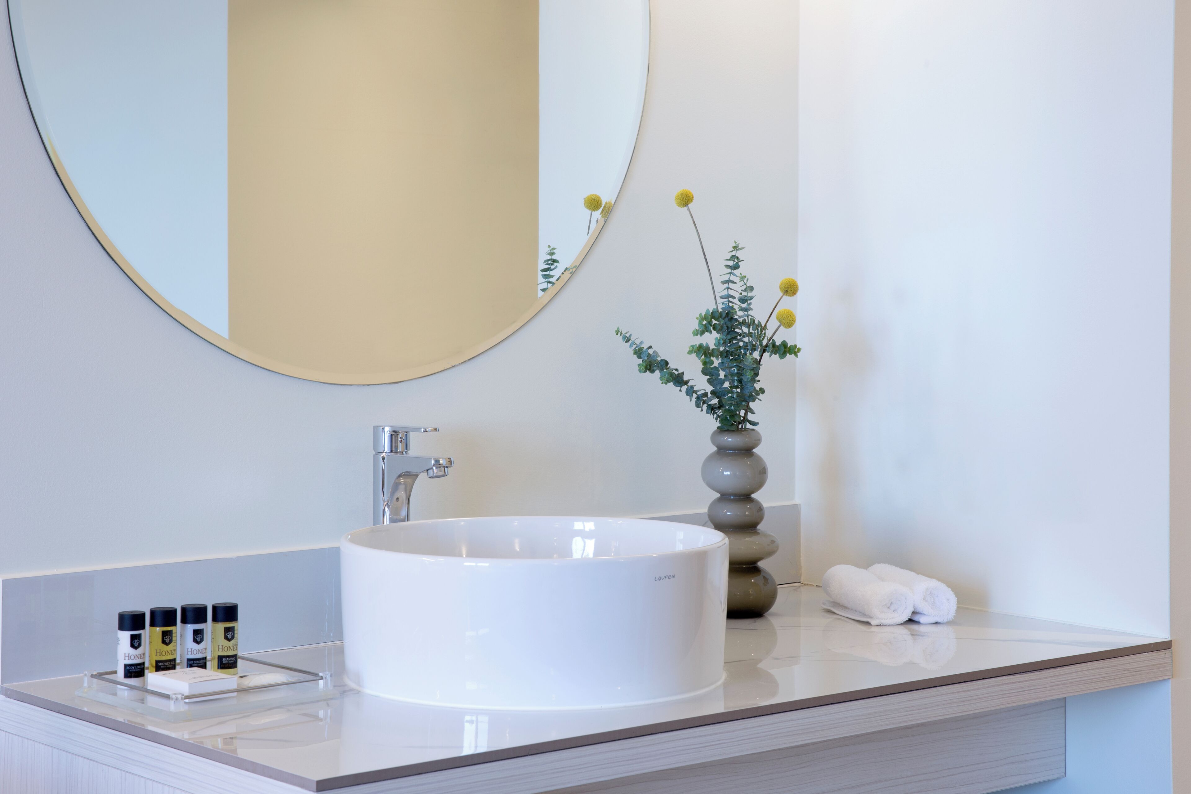 Un baño moderno con un lavamanos blanco y redondo, un espejo grande y un florero con flores amarillas.