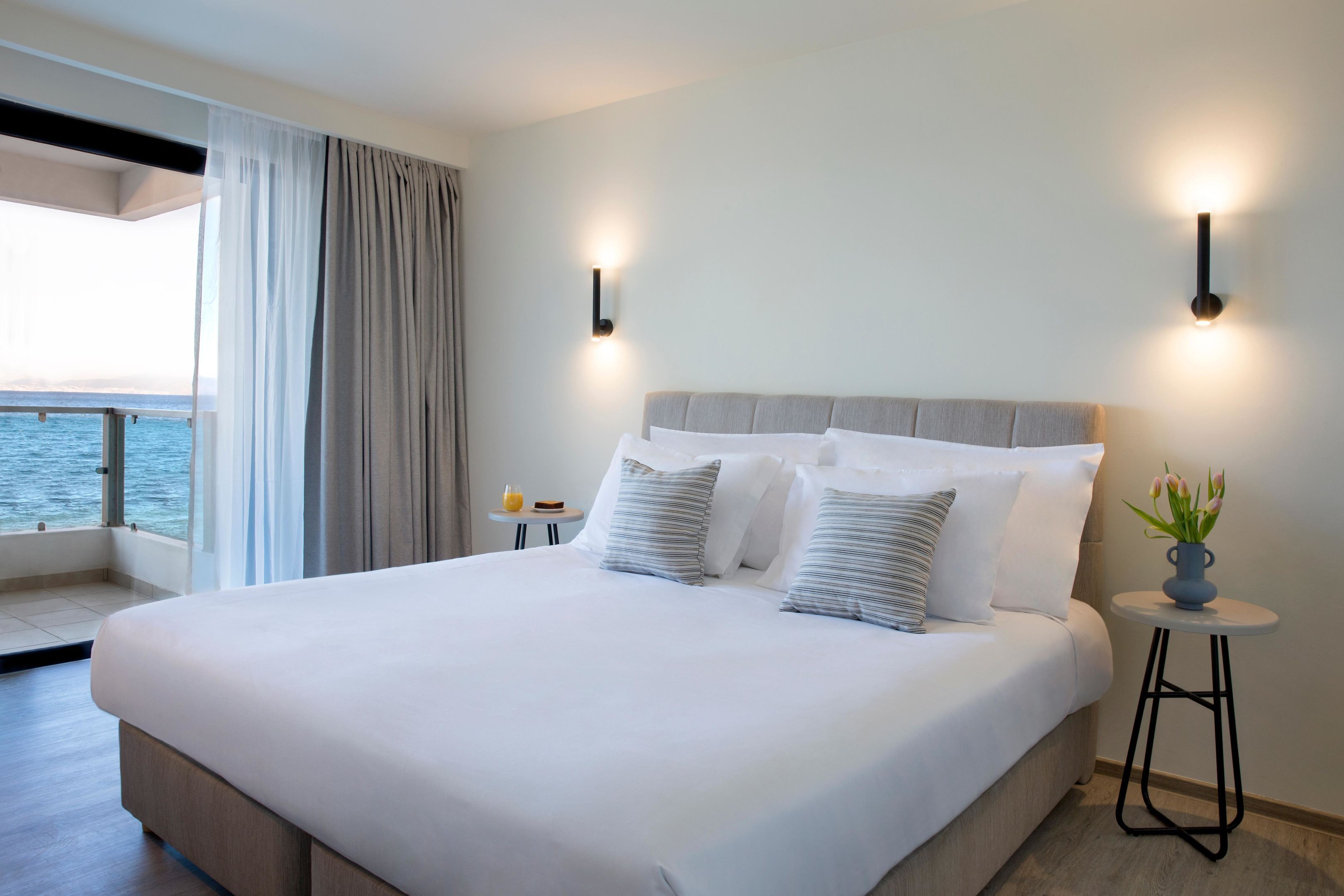 A hotel room with a large bed, two bedside tables, a small round table with a vase of flowers, and a view of the sea through a balcony.