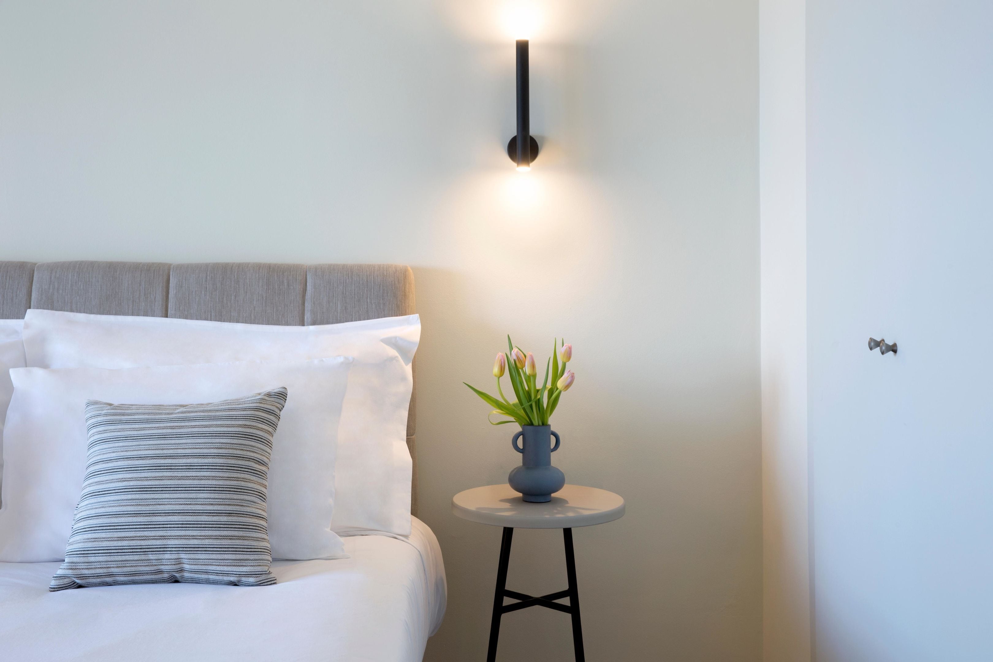 A neatly made bed with a striped pillow, a small round table with a vase of flowers, and a wall-mounted light fixture.