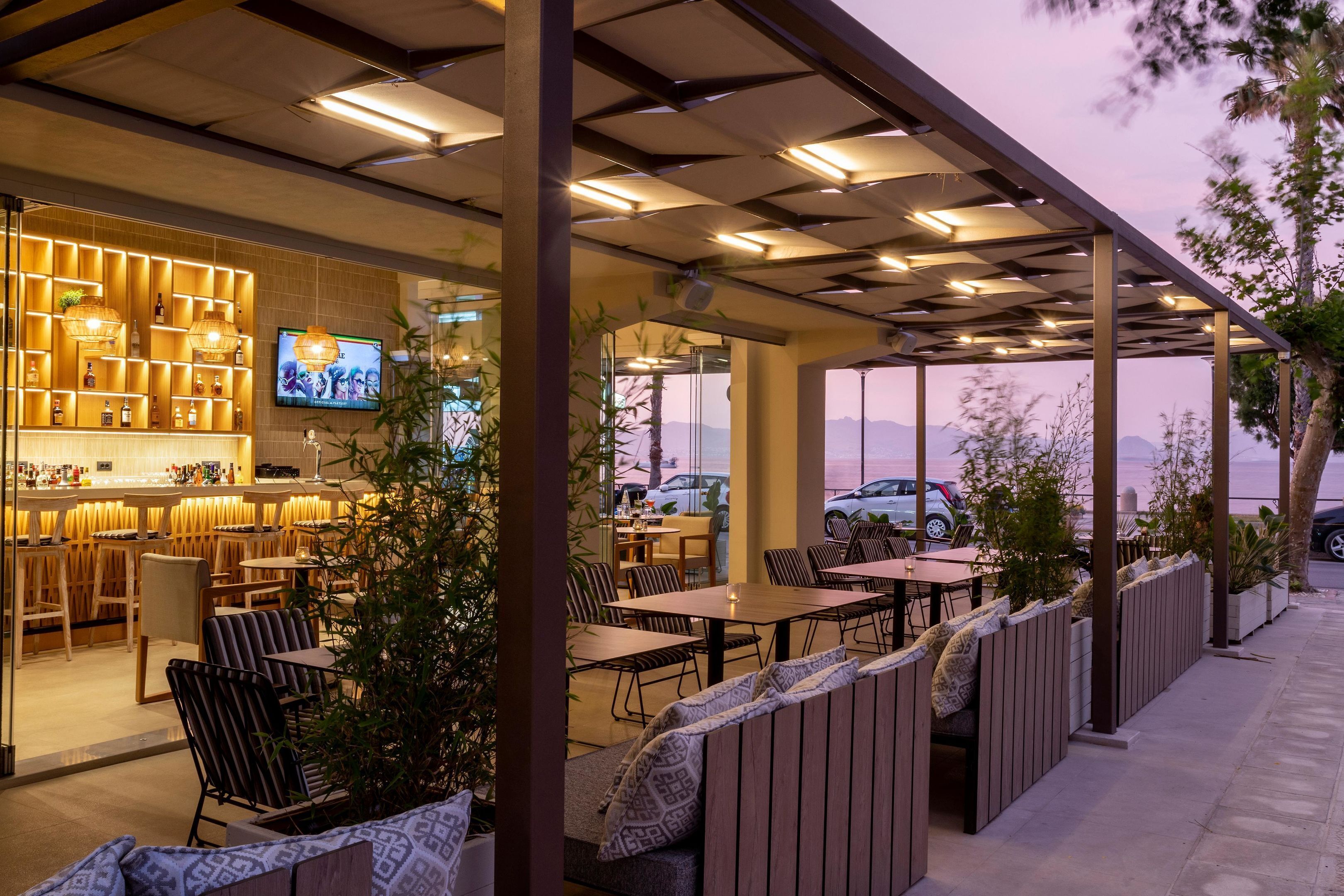 An outdoor restaurant with tables and chairs, a bar area with shelves of bottles, and a TV screen.