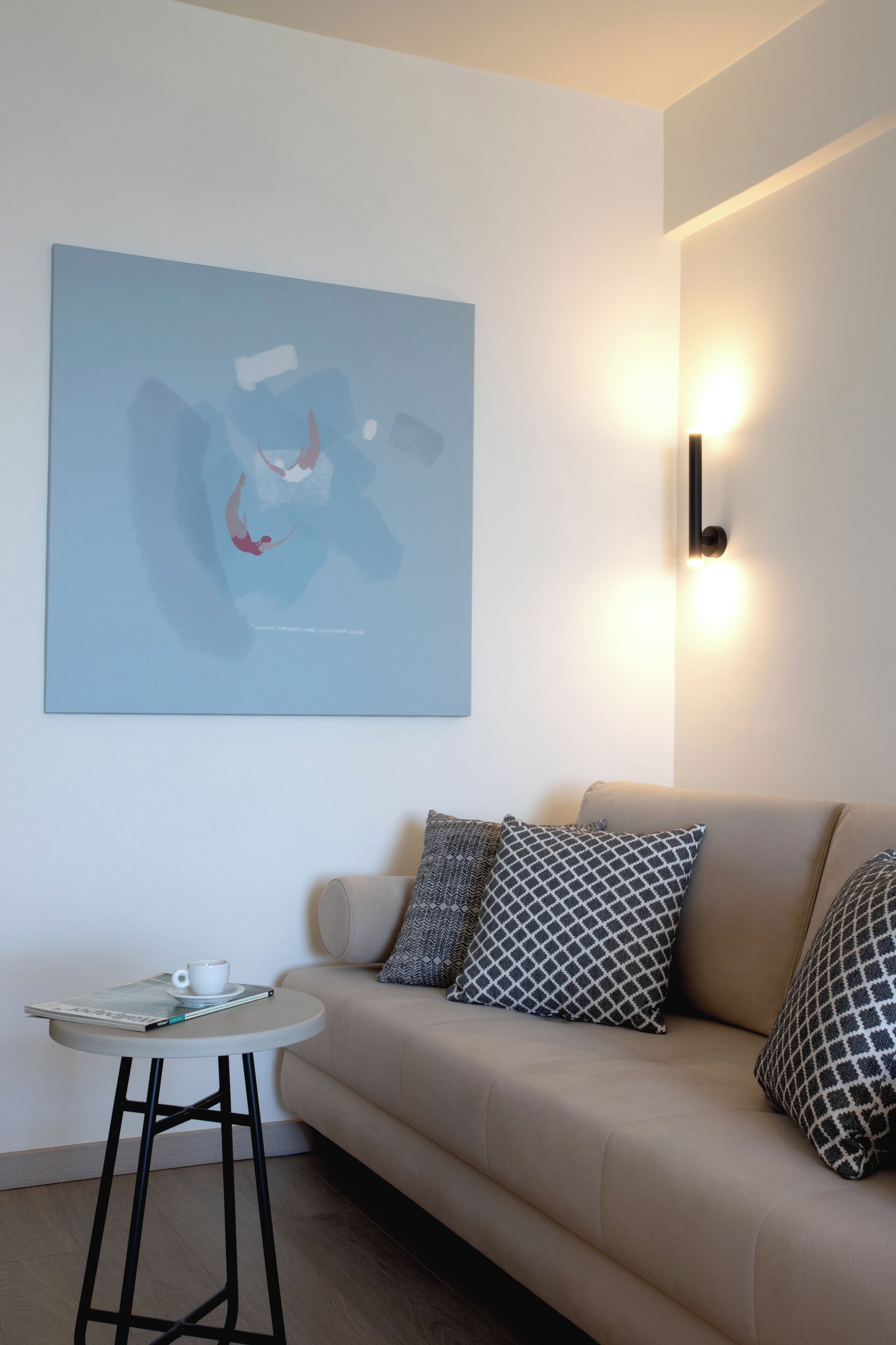 Una sala de estar moderna con un sofá color beige, cojines estampados, una pequeña mesa redonda con una taza y un libro, y un cuadro abstracto grande en la pared.