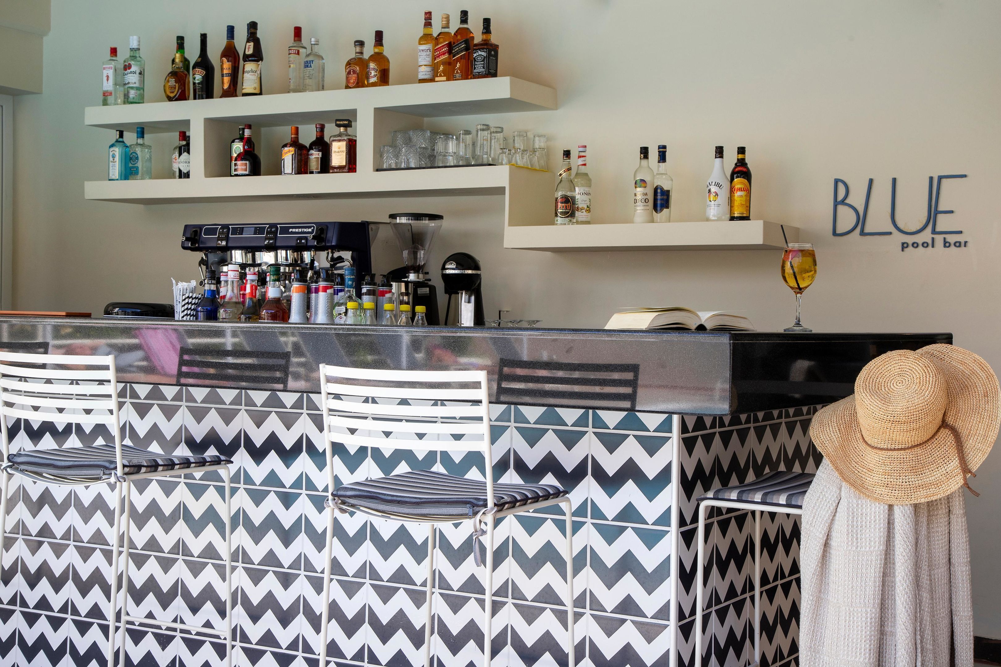 Una zona de bar con una pared de diseño en zigzag, estantes con botellas y vasos diversos, una máquina de café y un sombrero en una silla.