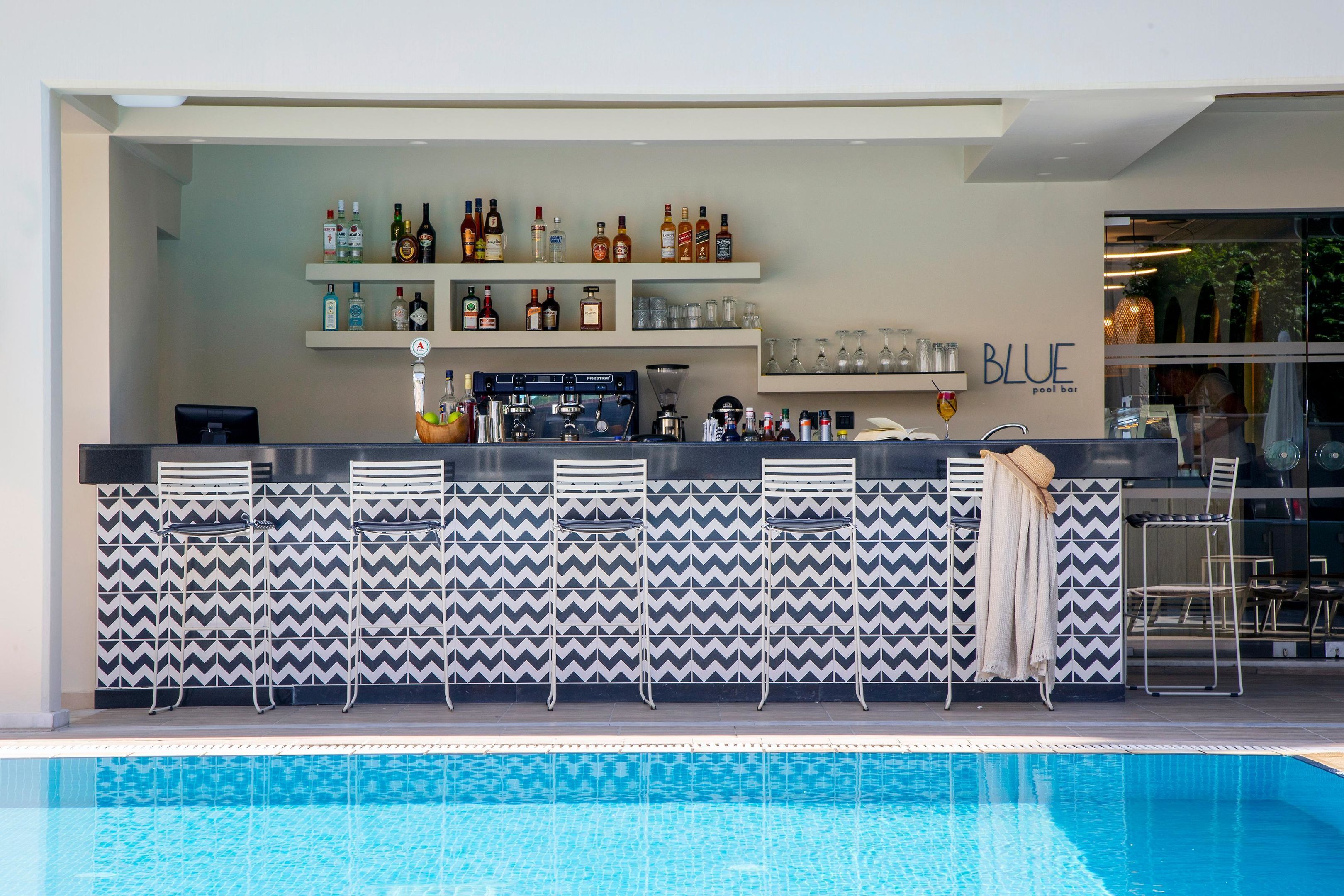 Un bar junto a la piscina con un muro de diseño en forma de tablero de ajedrez, taburetes y una selección de botellas y vasos en estantes.