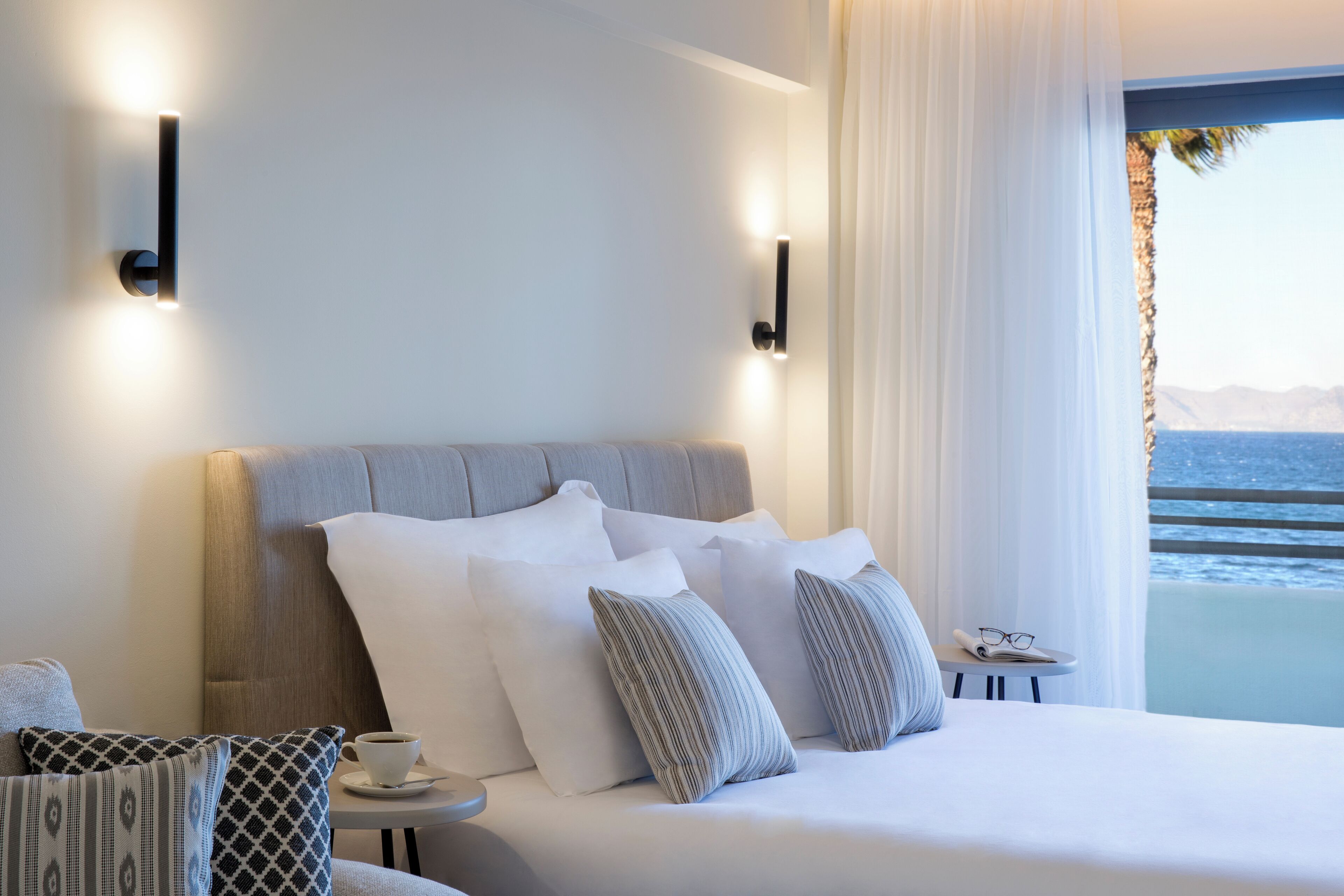 A modern hotel room with a large bed, a small table with a cup, and a view of the sea.