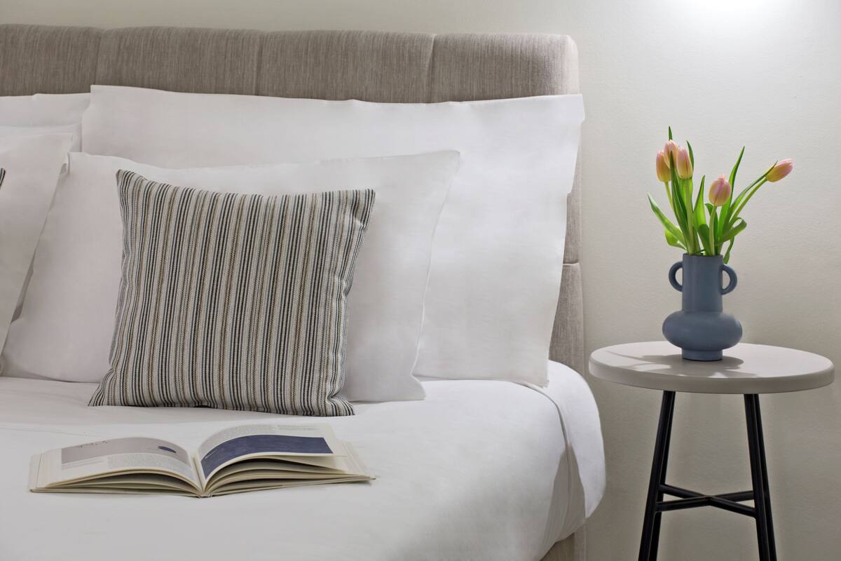 Een keurig opgemaakt bed met witte lakens en gestreepte kussens, een open boek op het bed, en een bijzettafeltje met een vaas tulpen.