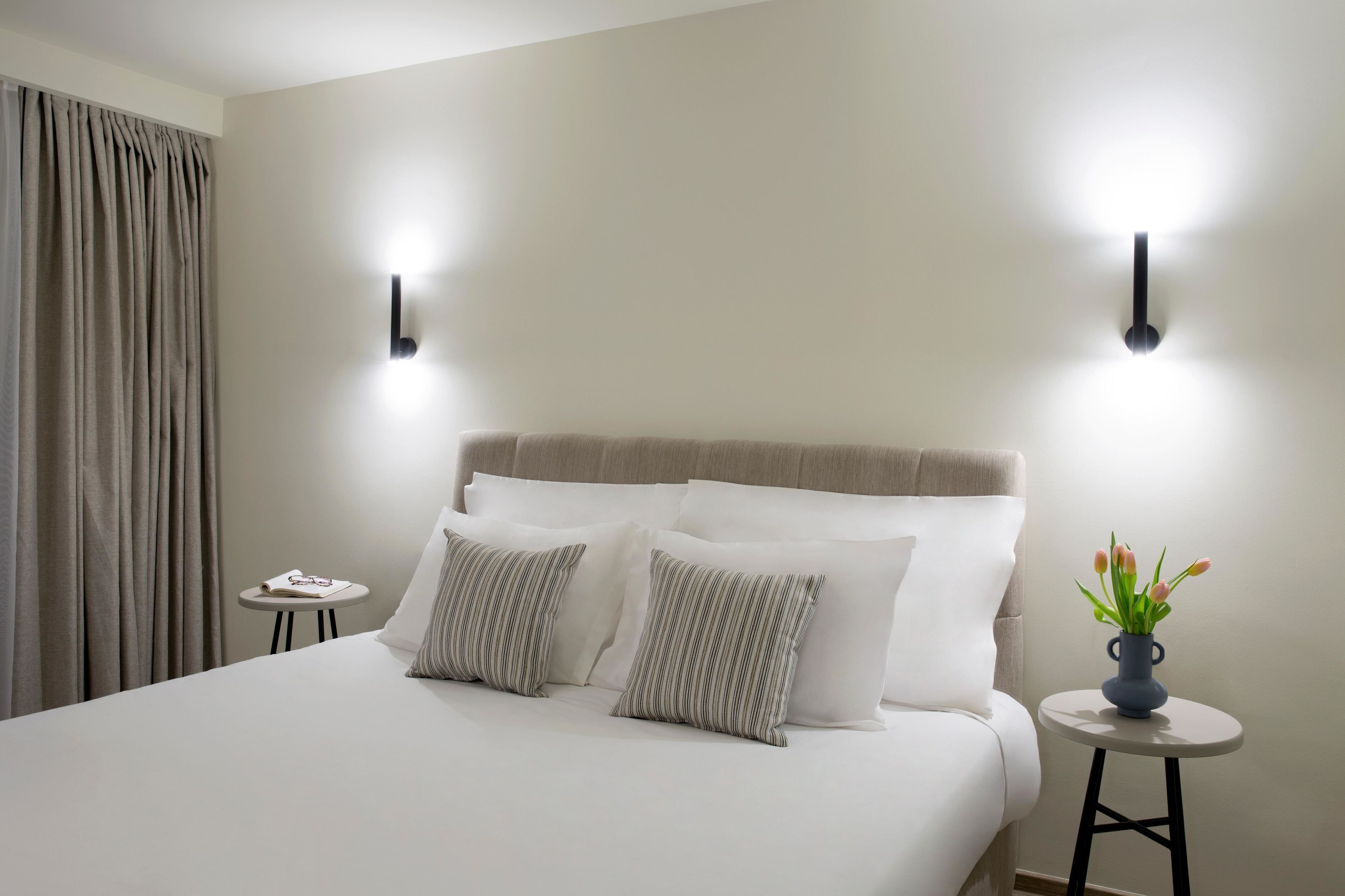 A neatly made bed with white linens and striped pillows, a small round table with a vase of flowers, and two wall-mounted lights.