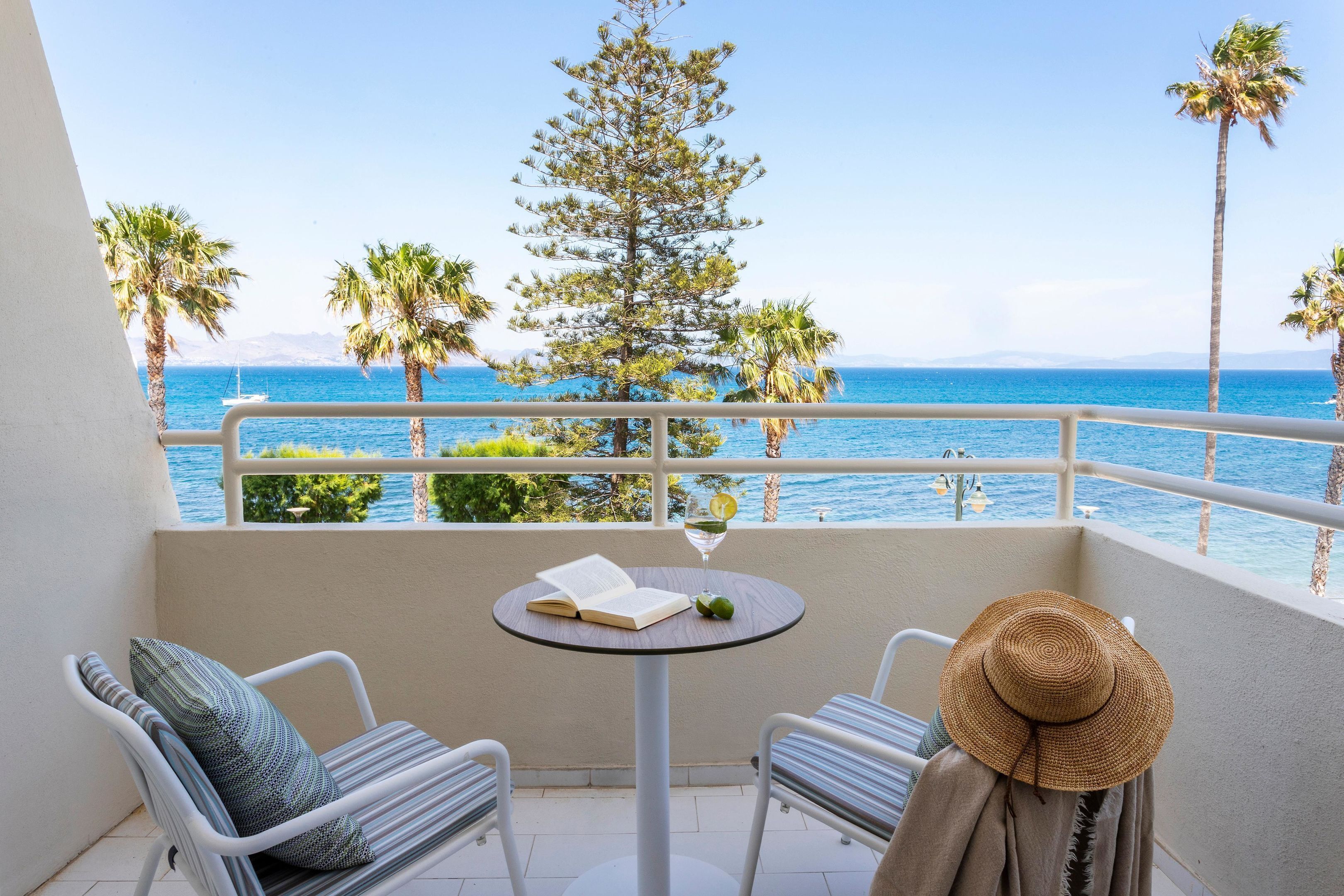 A balcony with a view of the sea, two chairs, a table with a book and a glass, and a straw hat.