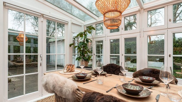 A dining area with a table set for four, surrounded by large windows and a wicker chair with a fur throw. - Chalet Kieftveen with hot tub for four Person (Voorthuizen)