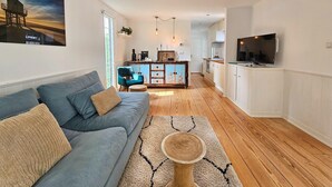 A modern living room with a grey sofa, a coffee table, and a kitchen in the background. - Chalet Kieftveen with hot tub for four Person (Voorthuizen)