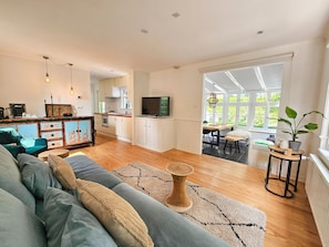 A spacious living room with a sofa, a coffee table, and a TV. There is a kitchen area with cabinets and a dining table.