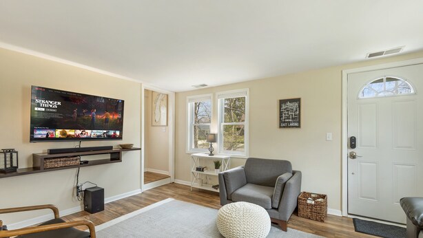 Una sala de estar con una televisión de pantalla plana, un sofá gris, un pouf redondo, una mesa de centro de madera y una estantería.