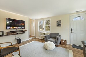A living room with a flat-screen TV, a grey sofa, a round pouf, and a wooden floor.