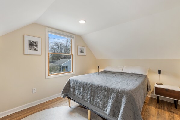 A bedroom with a sloped ceiling, a large window, a bed with a gray quilt, a bedside table with a lamp, and two framed pictures on the wall.