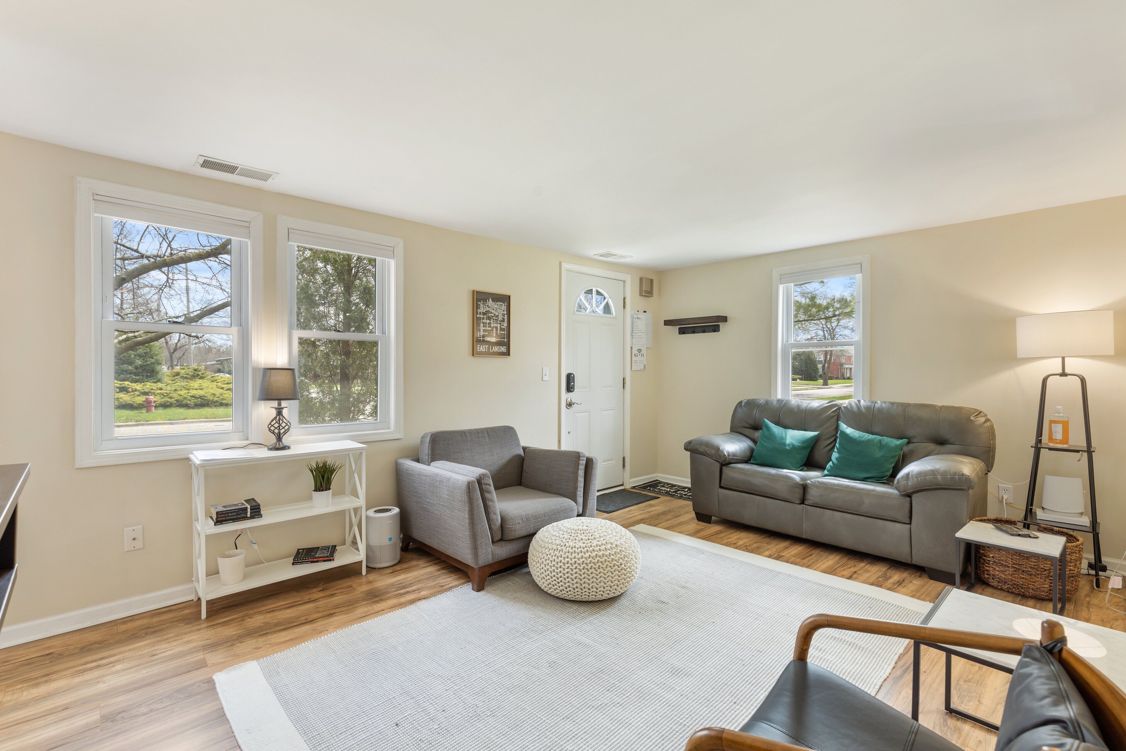 Ein Wohnzimmer mit grauem Sofa, grauem Sessel, weißem Konsolentisch und einem runden Pouf.