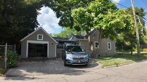Un garaje residencial con un coche aparcado, una casa con tejado inclinado y un árbol con flores blancas.