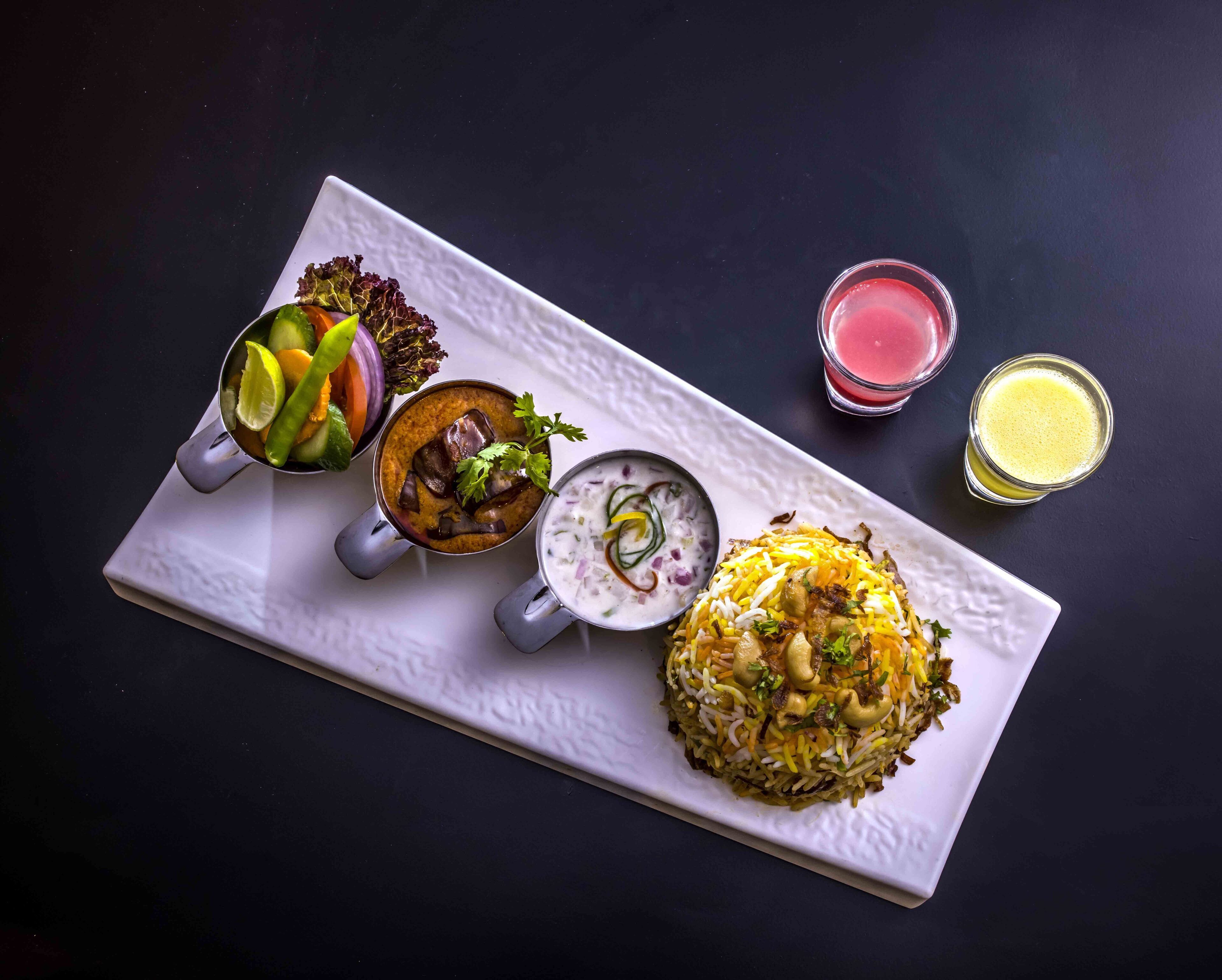 Een rechthoekige witte dienblad met diverse gerechten, waaronder een kom curry, een kom yoghurtgerecht, een bord biryani en twee glazen sap.