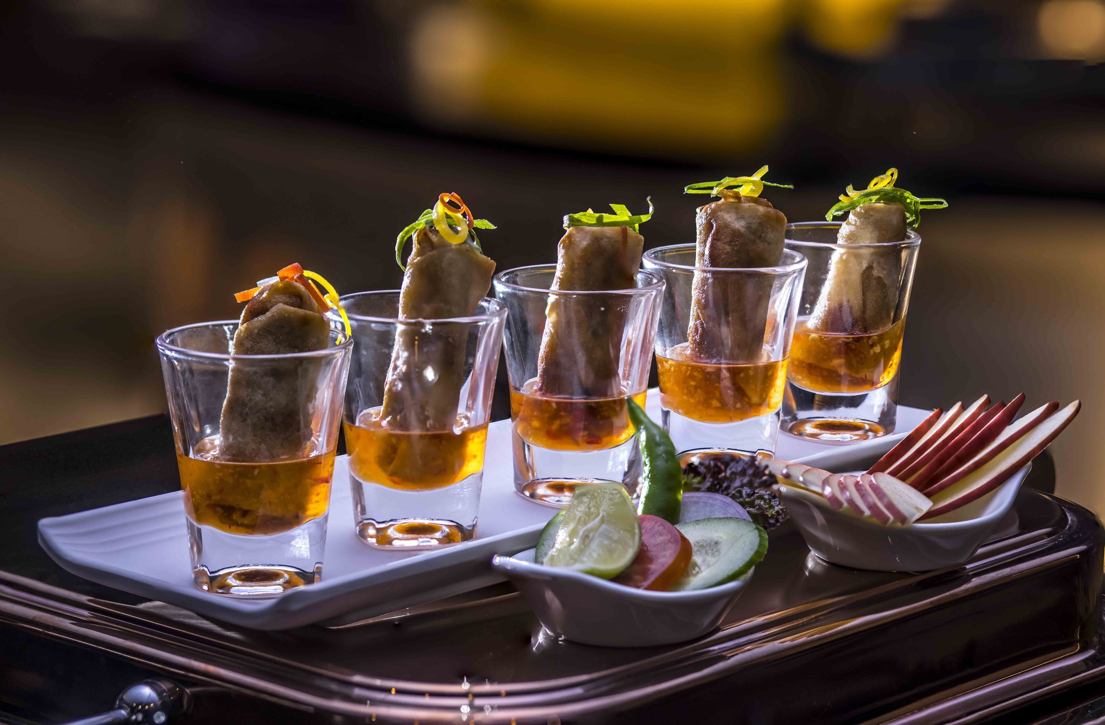 A tray with shot glasses containing a beverage and garnished with a slice of lime and cucumber, accompanied by a bowl of skewers and a small dish of olives.