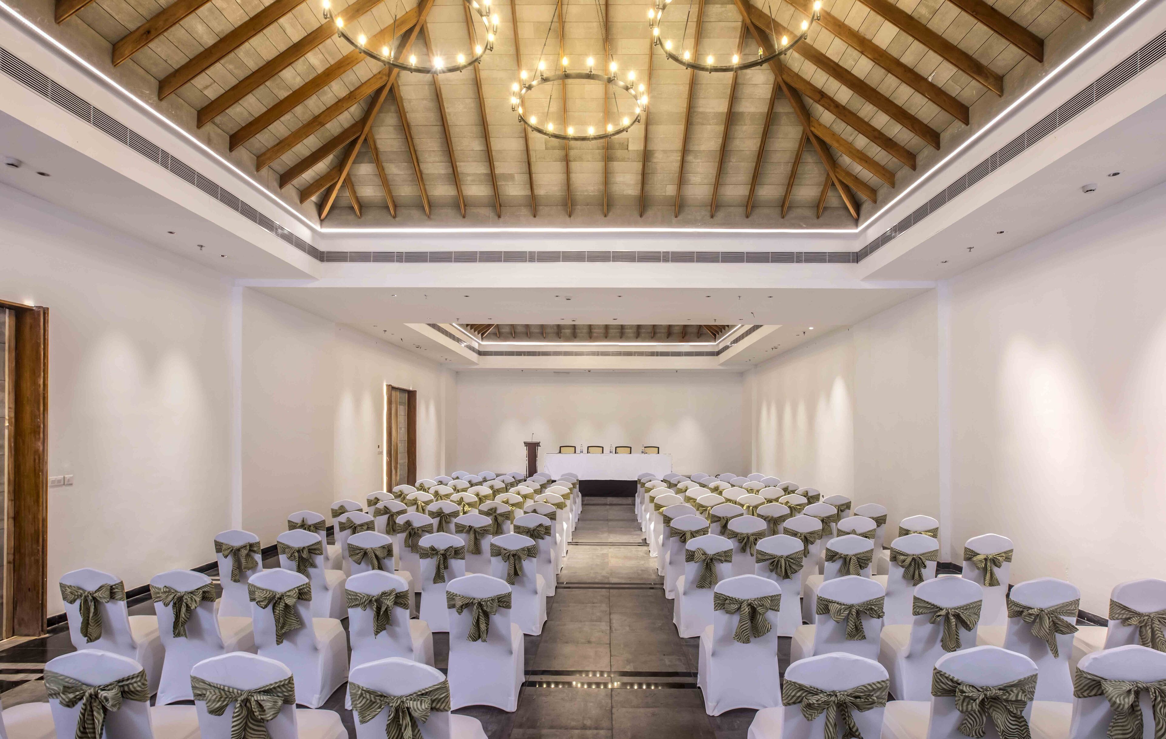 A conference room with rows of white chairs, a long table at the front, and a high wooden ceiling with chandeliers.