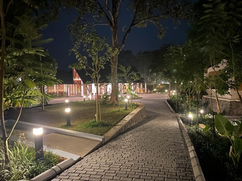 A nighttime pathway leading to a building with a red roof, surrounded by greenery and illuminated by pathway lights.