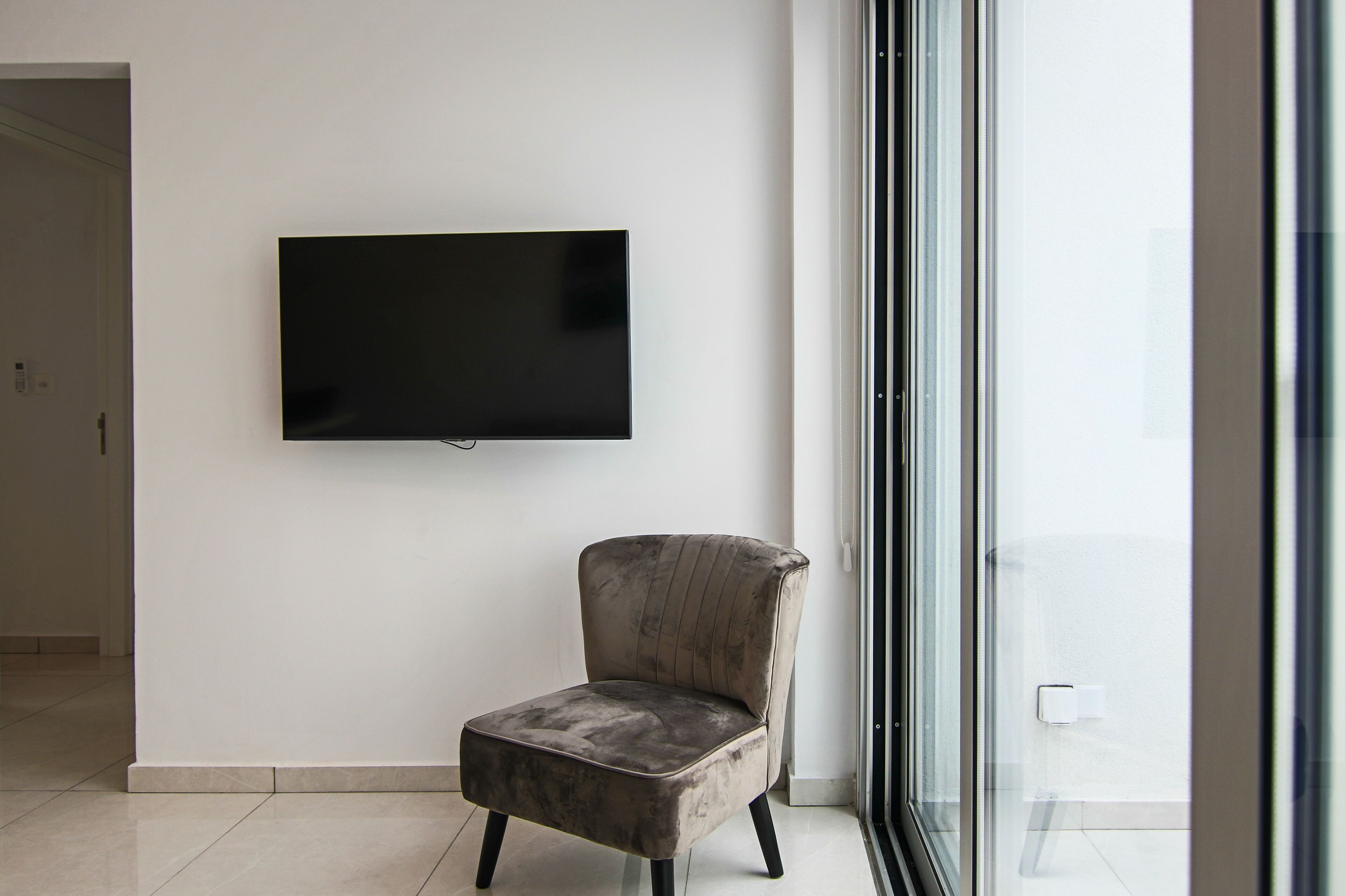 A modern living room with a flat-screen TV mounted on the wall, a plush armchair, and a large glass door leading to a balcony.