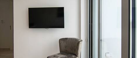 A modern living room with a flat-screen TV mounted on the wall, a plush armchair, and a large glass door leading to a balcony.