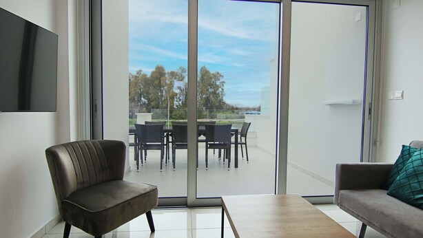 A modern living room with a sofa, a table, and a chair. There is a sliding glass door leading to an outdoor area with a table and chairs.