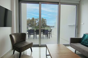 A modern living room with a sofa, a table, and a chair. There is a sliding glass door leading to an outdoor area with a table and chairs.