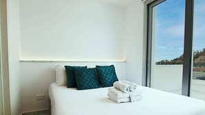 A neatly made bed with white and teal pillows, a folded towel, and a visible power outlet on the wall.