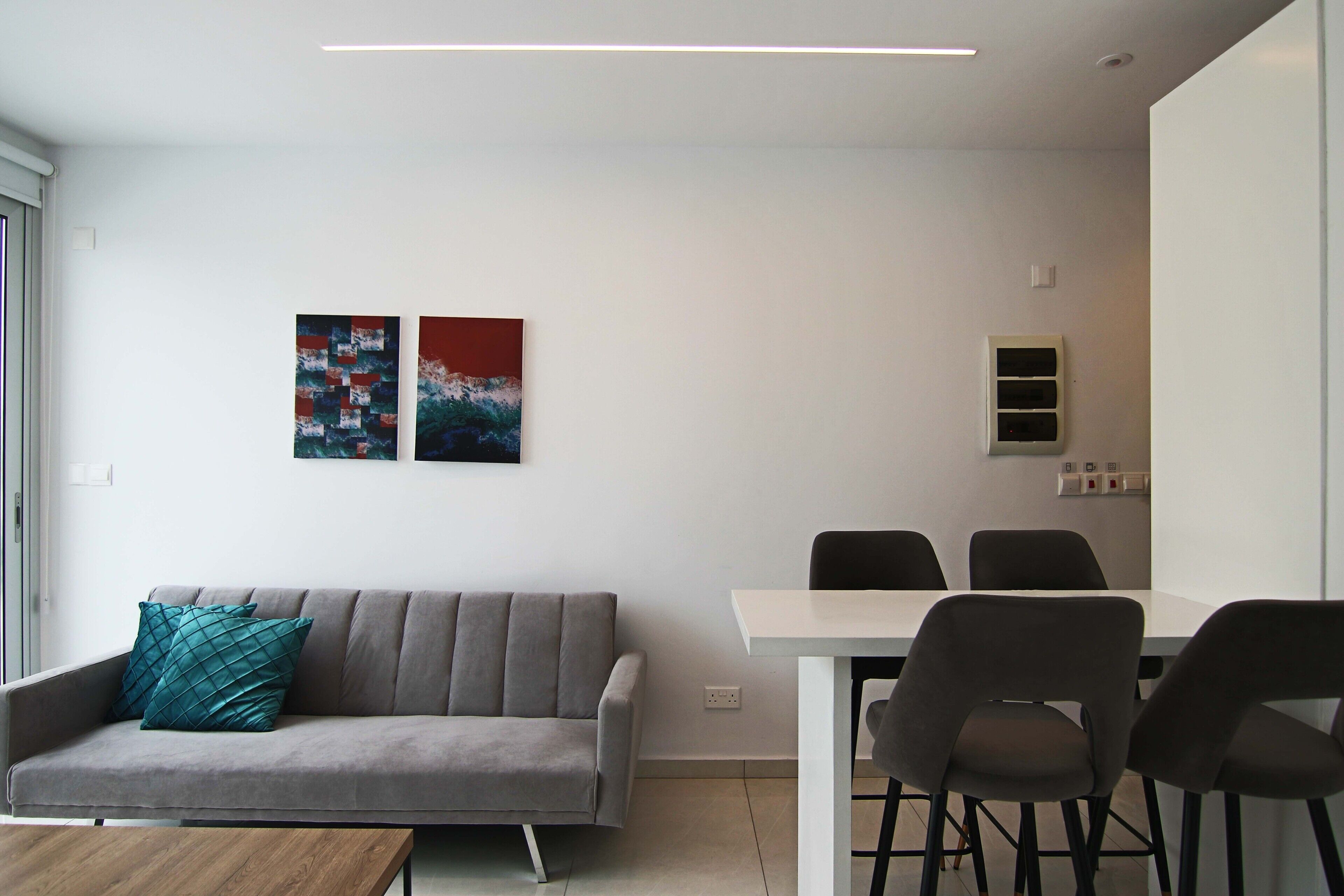 A modern living room with a grey sofa, teal pillows, a wooden coffee table, and a white dining table with four chairs. There are two abstract paintings on the wall.