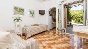 A cozy bedroom with a bed, a small table, and a chair. There is a kitchen area visible through an open door. - Effie Apartment in (Corfu)