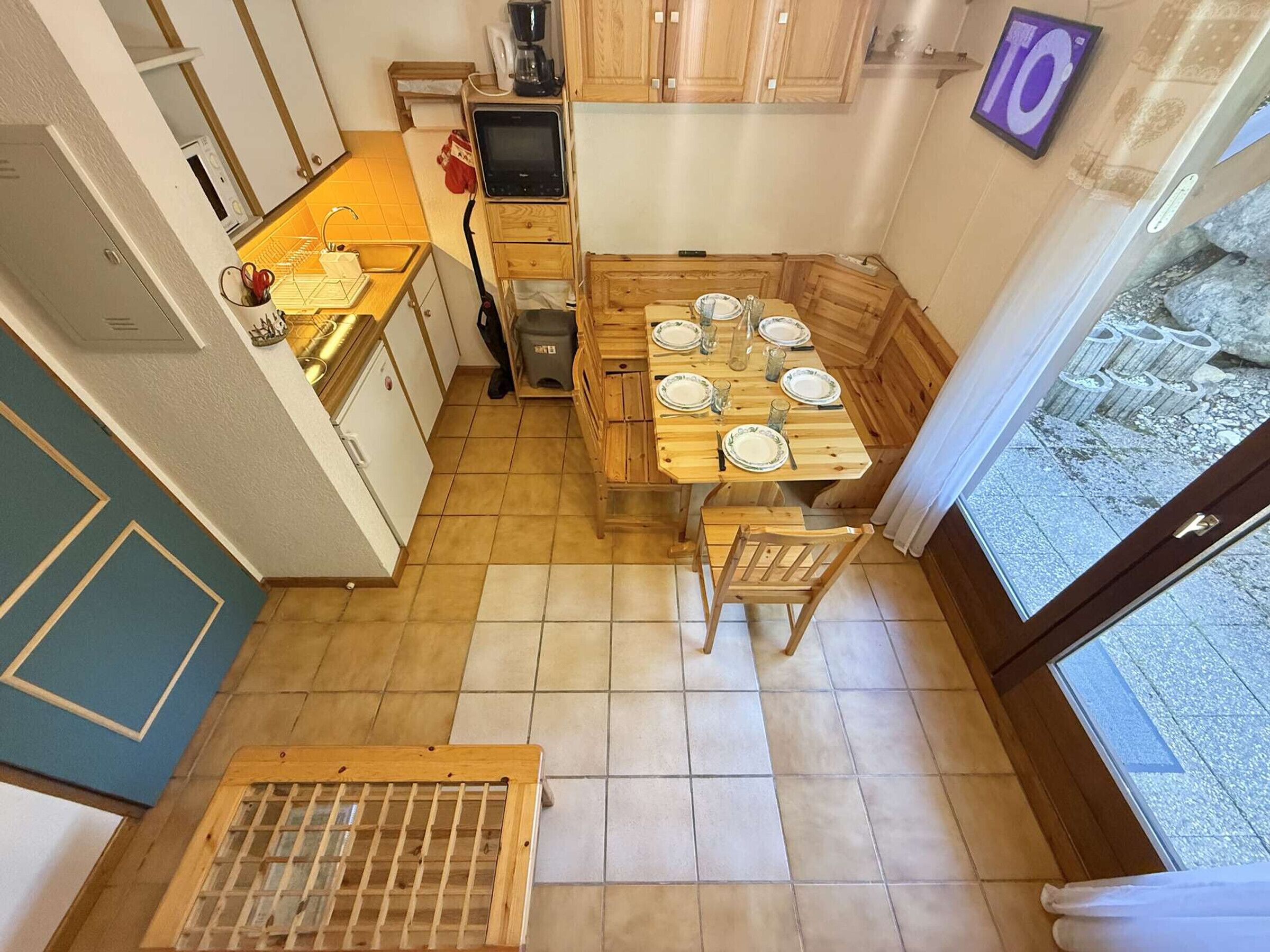 A compact kitchen with wooden cabinets, a dining table set for four, and a view of a rocky outdoor area through a glass door.