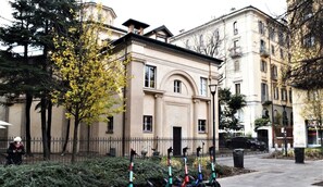 A historic building with a classical facade, a gated entrance, and a tree with yellow flowers.