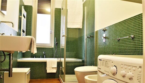 A bathroom with a green tiled wall, a white sink, a glass shower enclosure, and a washing machine.