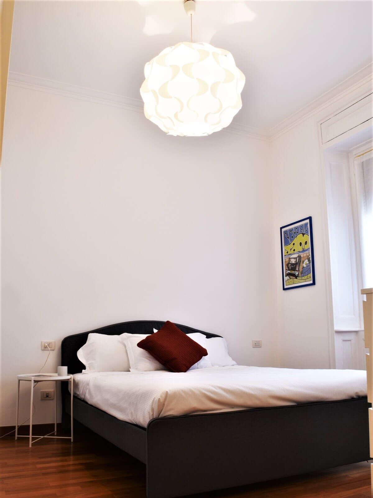Un dormitorio con una cama, una mesita de noche, una lámpara de pared e imagen enmarcada.