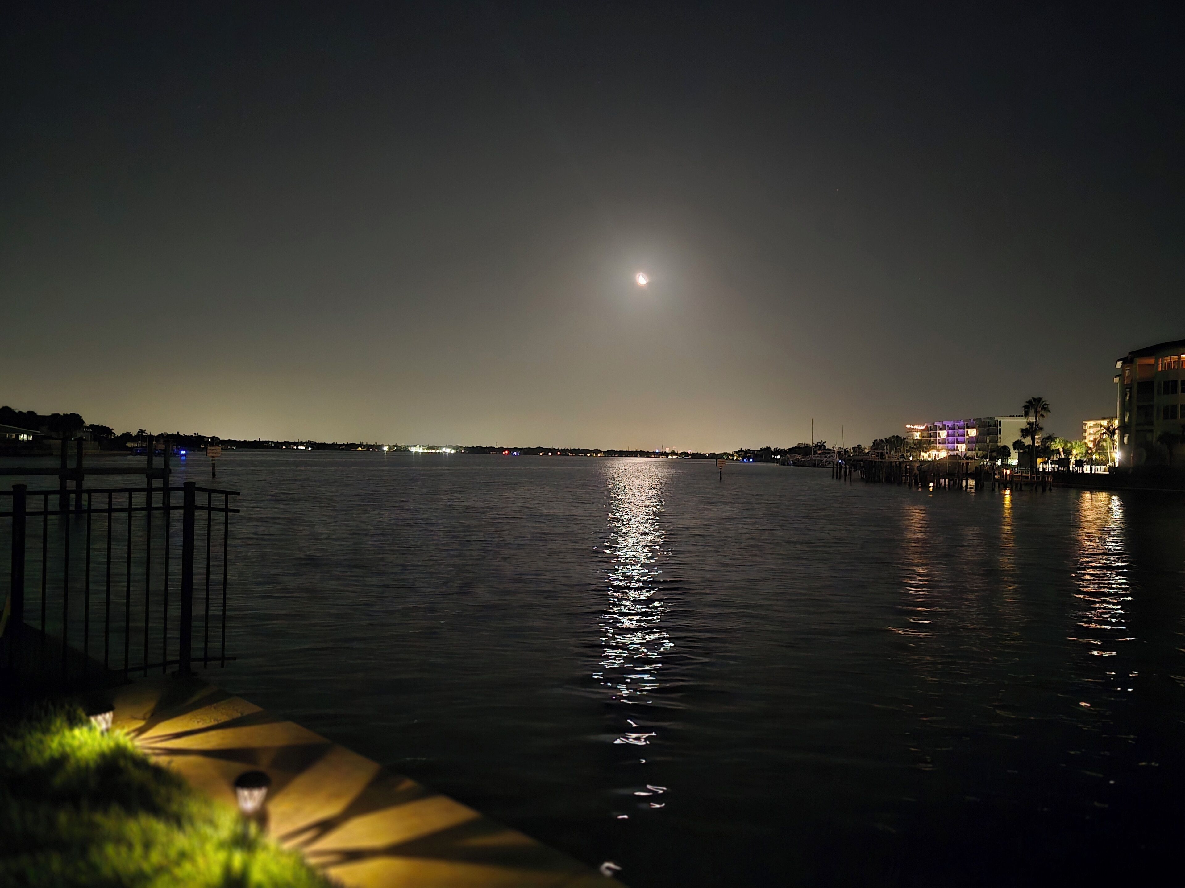 A nighttime waterfront scene with a calm body of water reflecting lights, a visible moon, and distant city lights.