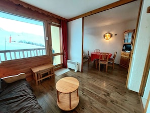 Um quarto com um banco de madeira, uma pequena mesa redonda, uma mesa de jantar com cadeiras, um radiador e uma televisão em um armário de madeira.