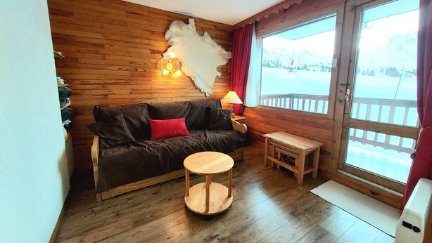 Ein gemütliches Zimmer mit Holzboden, einem braunen Ledersofa mit rotem Kissen, einem kleinen Holztisch und einer Lampe. Ein großes Fenster bietet Blick auf eine verschneite Landschaft.