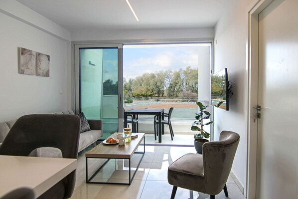 A modern living room with a sofa, a coffee table, and a dining area. There is a large window offering a view of trees and a tennis court.