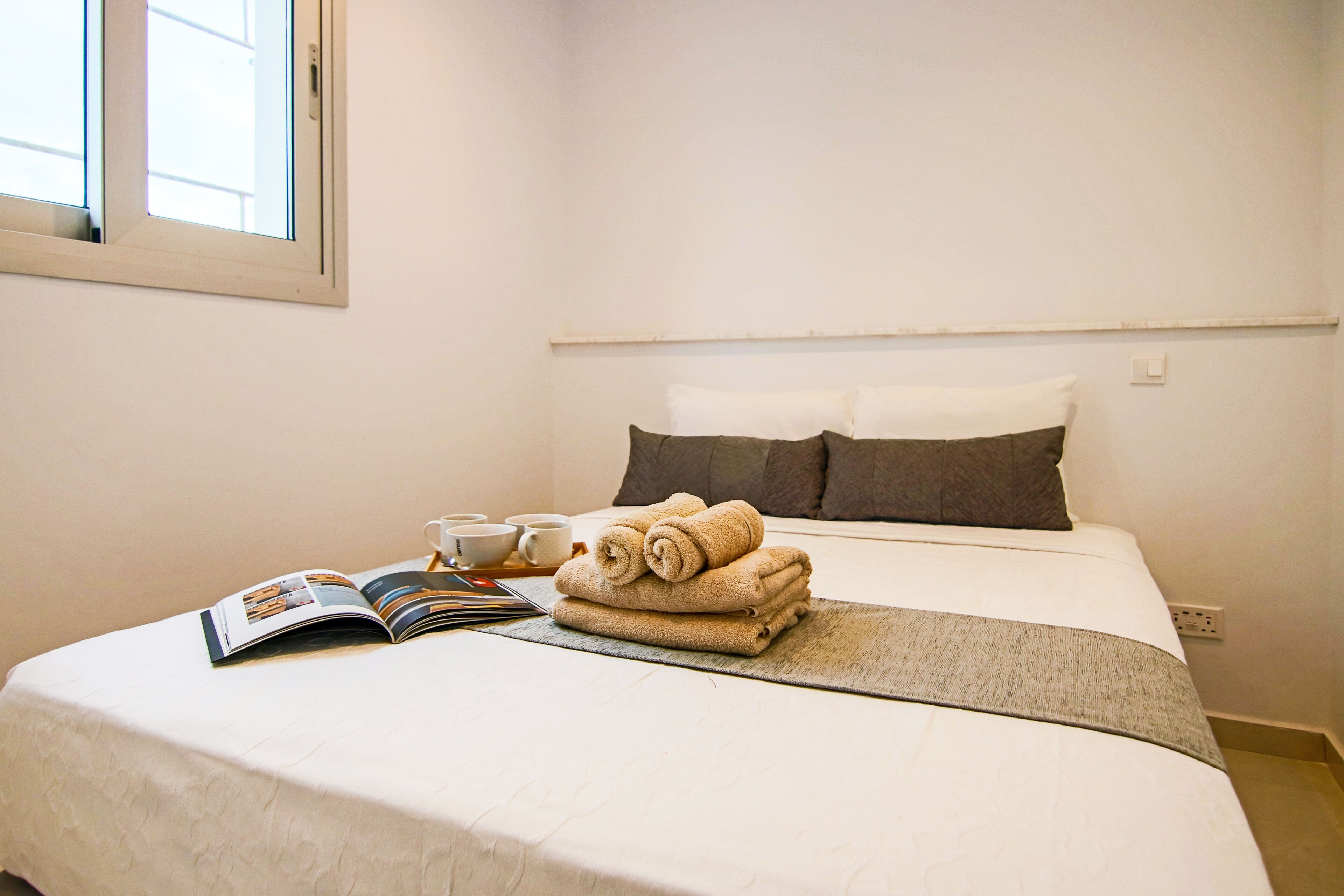 A neatly made bed with a folded towel, a magazine, and a cup of coffee on the bedside table.