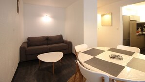Une petite pièce chaleureuse et accueillante, avec un canapé marron, une table blanche ornée d’un chemin de table à carreaux, et un coin cuisine comprenant un réfrigérateur et une cuisinière.