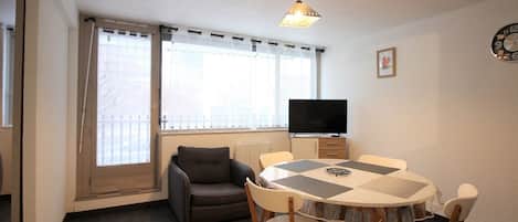 Um quarto pequeno e bem iluminado com uma mesa redonda, quatro cadeiras, um sofá, uma televisão e uma janela com cortinas.