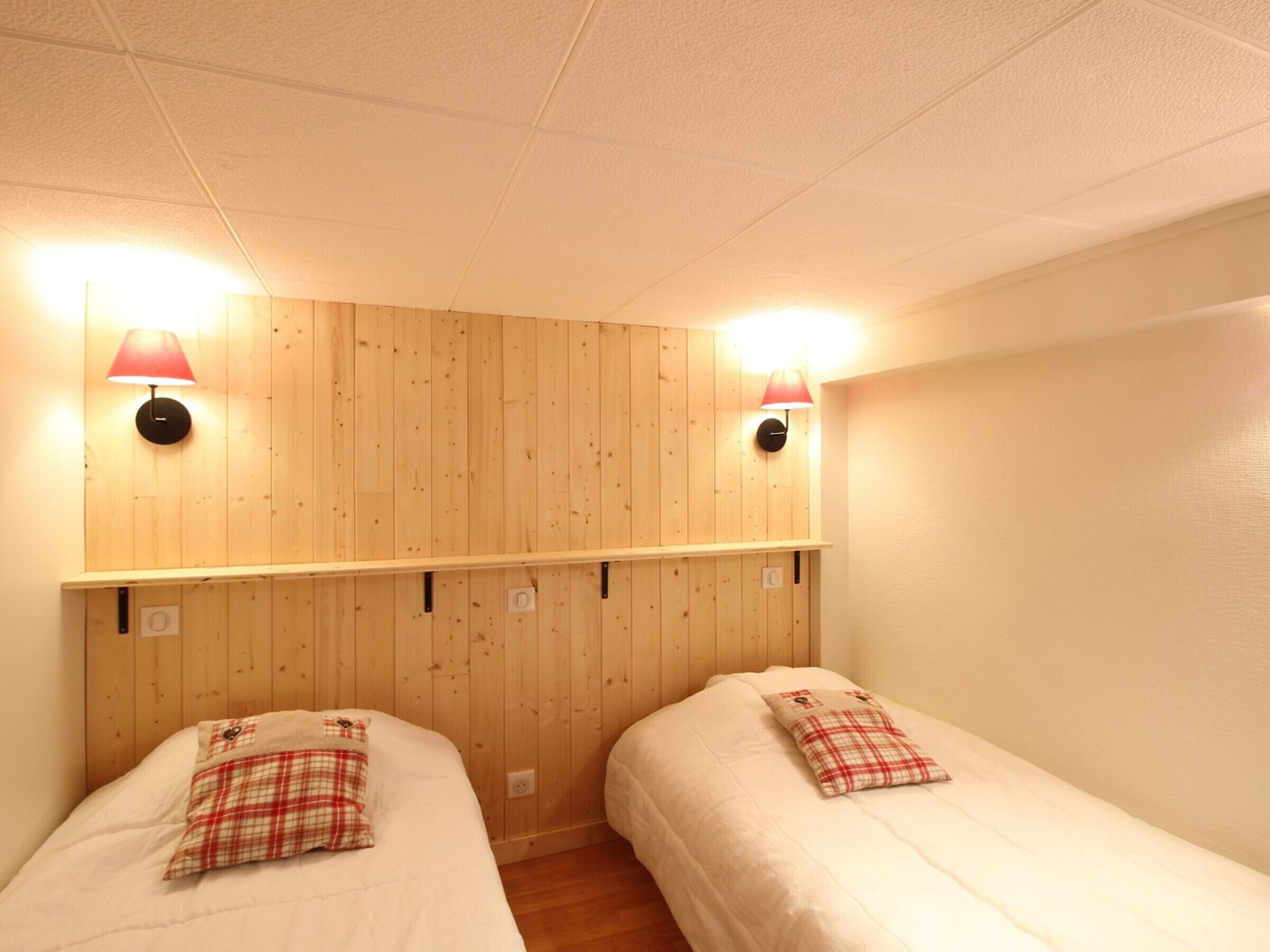 A room with two beds, wooden panel walls, and two wall-mounted lamps.