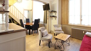 A well-lit living room with a dining area, a staircase, a TV mounted on the wall, and a large window. - Cozy T2 bis with Wi-Fi, 100m from center, sleeps 6, ski & bike storage (Mont-Dore)