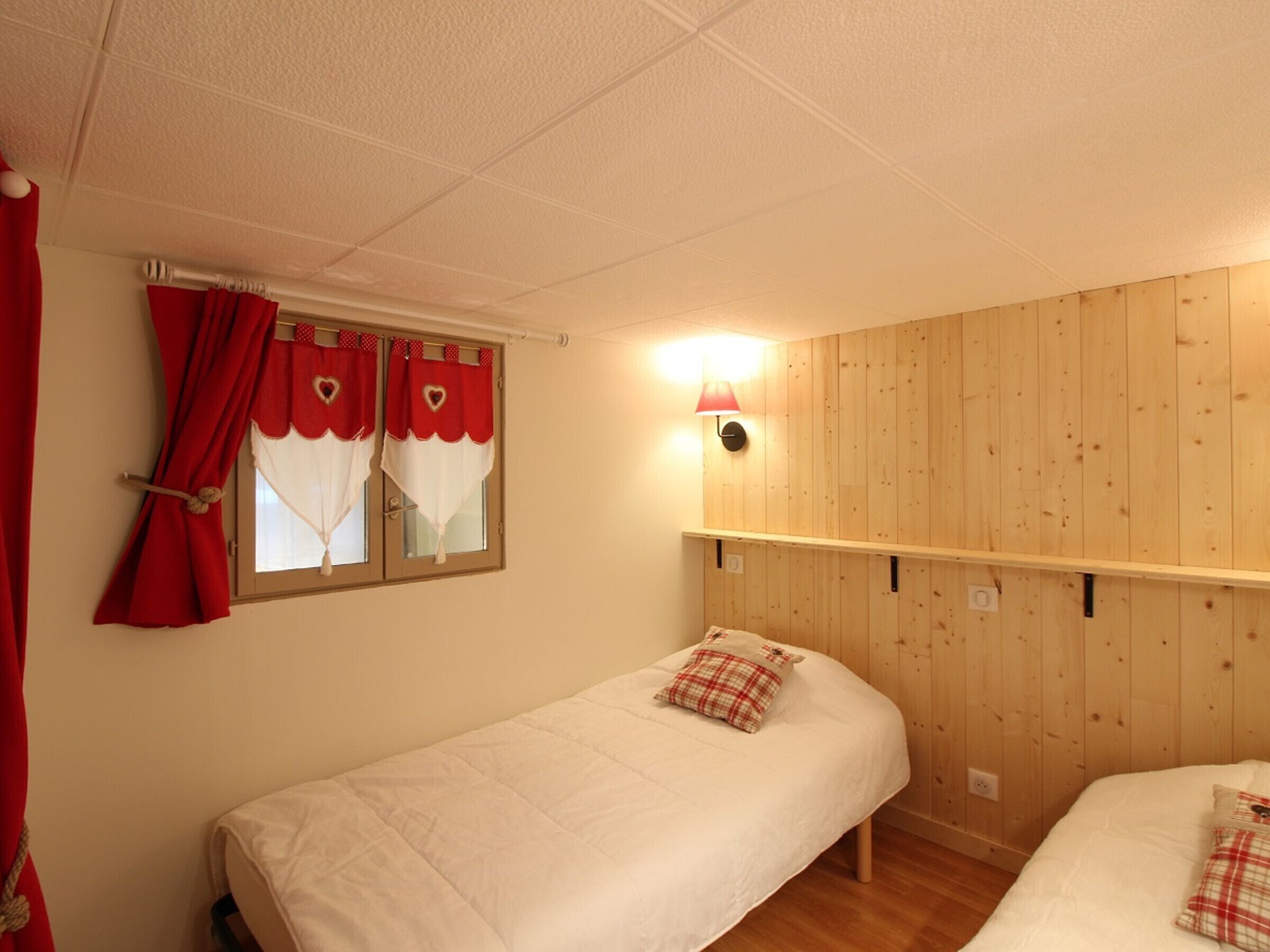Un dormitorio con dos camas individuales, una ventana con cortinas rojas, un estante fijado a la pared y una lámpara de pared.