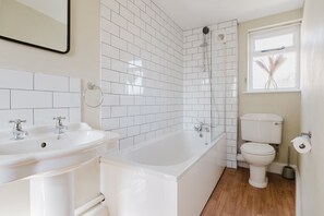 A modern bathroom with white tiled walls, a bathtub, two sinks, a toilet, and a window with a view of palm trees. - A beautifully presented two bedroom Cotswold stone cottage in the Cotswolds - Cobblers Cottage, More (Moreton-in-Marsh)