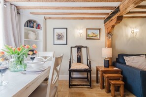 A cozy living room with a dining area, featuring a wooden chair, a blue sofa, a small wooden table, and a lamp. - A beautifully presented two bedroom Cotswold stone cottage in the Cotswolds - Cobblers Cottage, More (Moreton-in-Marsh)