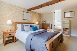 A bedroom with a stone wall, a wooden bed frame, a nightstand with a lamp, a dresser, and a window with blinds. - A beautifully presented two bedroom Cotswold stone cottage in the Cotswolds - Cobblers Cottage, More (Moreton-in-Marsh)