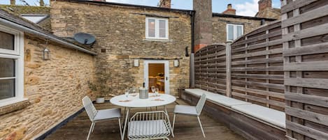 A stone building with a wooden fence, a round table with chairs, and a door leading to an interior space.